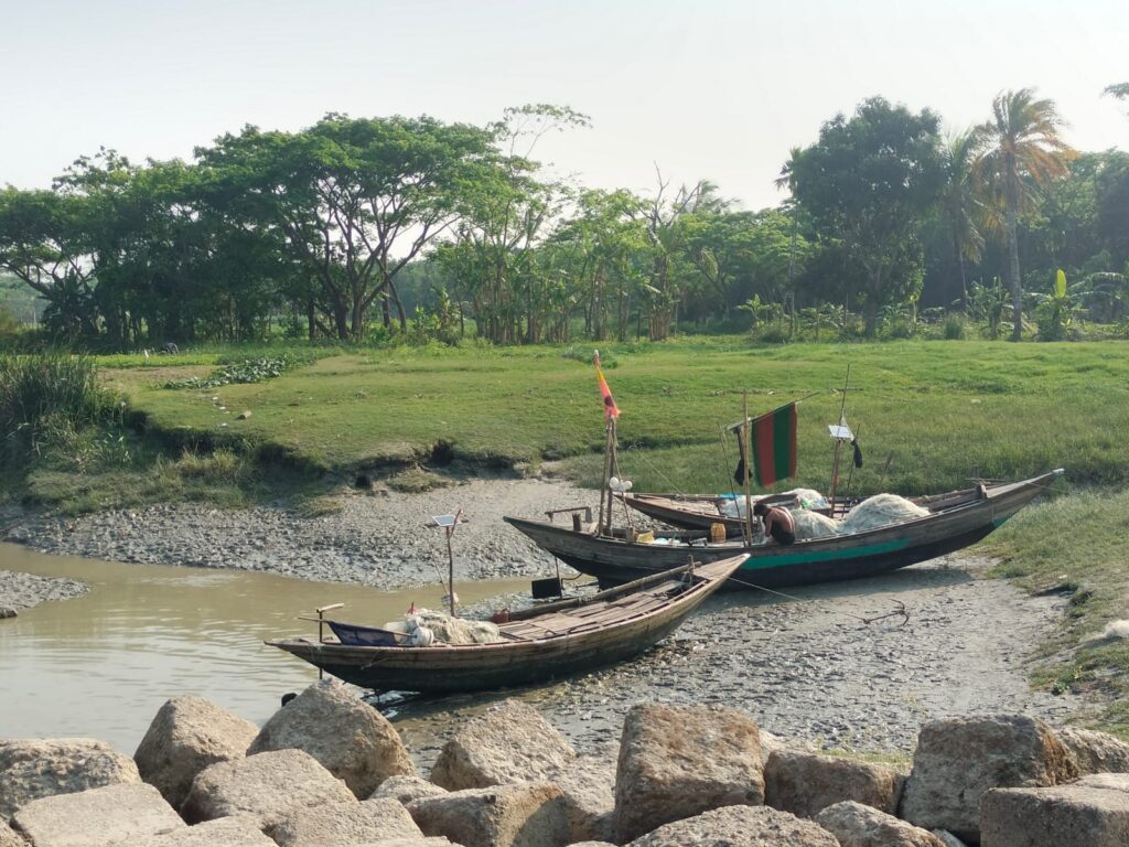 নদীর তীরে নোঙর করা মাছ ধরার নৌকা, সবুজ প্রকৃতি আর বাংলার গ্রামীণ জীবনের চিরচেনা সৌন্দর্য।